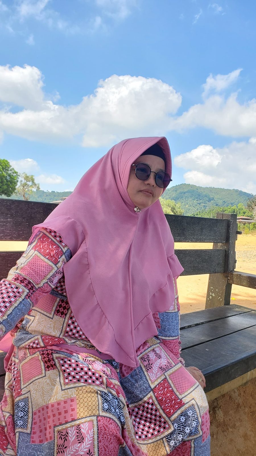 Ummi berkacamata hitam duduk di bangku dengan latar bukit dan pantai
